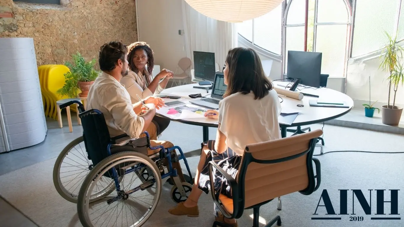 image de trois personnes autour d un bureau avec une personne en fauteuil roulant et le logo AINH 