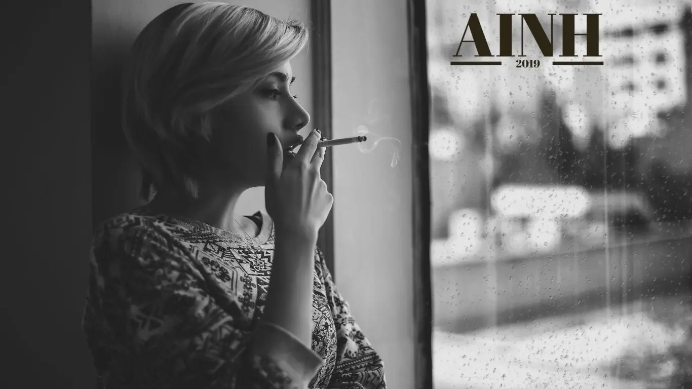 image en noir et blanc d une femme qui fume en regardant a travers la fenetre et il pleut et le logo AINH